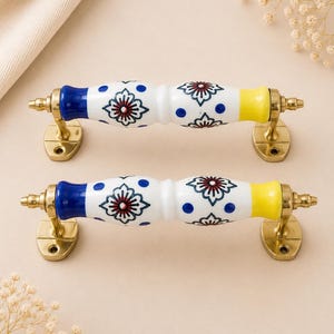 Op de afbeelding: Twee kastgrepen met een cilindrisch keramisch middendeel op een witte basis, voorzien van blauwe stippen en een rood-blauw bloemmotief. De uiteinden zijn afgewerkt in felblauw en geel, ondersteund door gepolijste messing steunen en bevestigingen.