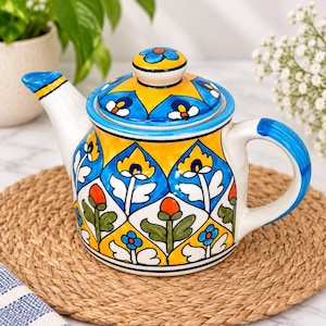 Op de afbeelding: Een keramische theepot met een handgeschilderd patroon van gele en blauwe ruiten met bloemmotieven in oranje, groen en wit. De theepot heeft een bijpassend deksel met een blauw bloemmotief en een gebogen handvat.