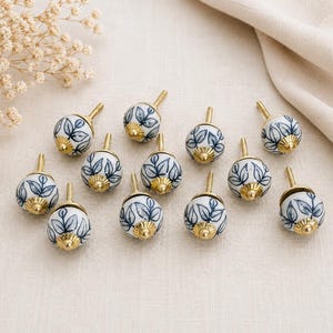 May include: A collection of eleven round ceramic knobs featuring a hand-painted blue leaf pattern on a white background. Each knob is finished with a metallic gold-toned base and a central screw attachment, arranged in a group on a light surface.