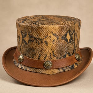 May include: A top hat featuring a tan and black snakeskin pattern on the crown. The hat includes a brown leather band around the base, decorated with three circular brass-toned metal studs, each embossed with a horse head profile design.