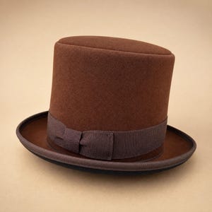 May include: A brown top hat with a flat crown and a structured brim. The hat features a dark brown grosgrain ribbon band wrapped around the base of the crown, which includes a small, centred bow detail. The material has a smooth, felted texture.