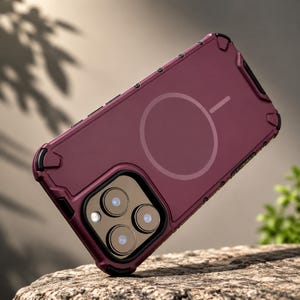May include: A deep burgundy smartphone case featuring a reinforced frame with black corner accents and a circular magnetic charging ring on the back. The case includes a square camera cutout with three circular lens openings and a flash aperture.