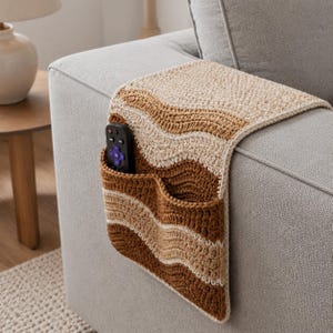 May include: A crocheted sofa armrest organizer featuring a wavy pattern in shades of cream, tan, and dark brown. The textile accessory drapes over the arm of a gray couch and includes a pocket containing a black remote control with purple buttons.