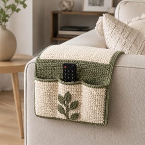 May include: A rectangular crochet sofa armrest organizer featuring a cream-colored base with olive green trim and a central leaf-shaped appliqué. The organizer includes three storage pockets, one of which holds a black rectangular remote control.