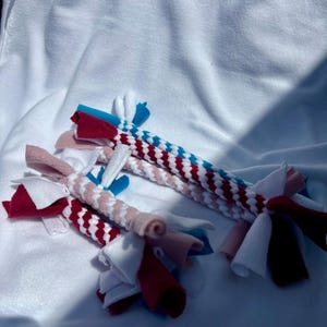 Könnte beinhalten: Zwei geflochtene Stoff-Zerrspielzeuge aus Fleece-Streifen. Ein Spielzeug hat ein Muster in Rot, Weiß und Blau, während das andere Farbtöne in Rosa, Weiß und Rot kombiniert. Beide haben verknotete Enden mit abstehenden losen Stoffstreifen.