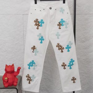 Op de afbeelding: Witte denim jeans versierd met kruisvormige patches. De kruisen hebben lichtblauwe, turquoise en luipaardprints. De jeans hangen aan een zwarte hanger.