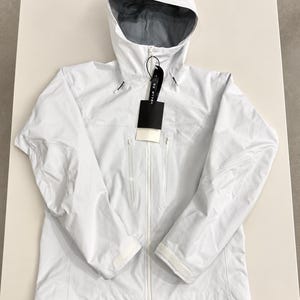 Può includere: Una giacca bianca con cappuccio dotata di cerniera frontale, due tasche verticali sul petto con chiusura a zip e polsini regolabili con velcro. La fodera interna è grigia e un'etichetta nera è fissata nella zona del colletto.