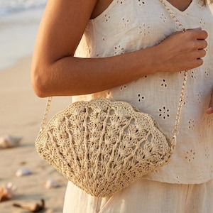 Könnte beinhalten: Beige, muschelförmige Umhängetasche mit goldener Kette. Die Tasche besteht aus gewebtem Material und hat einen gewellten Rand. Die Frau trägt ein weißes Oberteil und steht an einem Sandstrand.