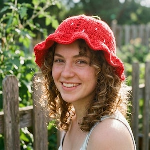 Op de afbeelding: Een felrode gehaakte bucket hat met een golvende, geplooide rand. De hoed is voorzien van kleine, gelijkmatig verdeelde witte geborduurde steken over de kroon, die het uiterlijk van een aardbei nabootsen met een klein groen steeltje bovenop.