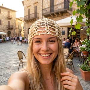 May include: A woman with long blonde hair smiles, wearing a cream-colored, open-weave head covering with gold sequins. The background shows a European town square with stone buildings and people.