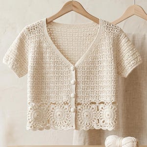 May include: A cream-colored short-sleeved crochet cardigan featuring a V-neckline, four round buttons down the center front, and a detailed circular floral pattern along the bottom hem, displayed on a wooden hanger on a gold metal rack.