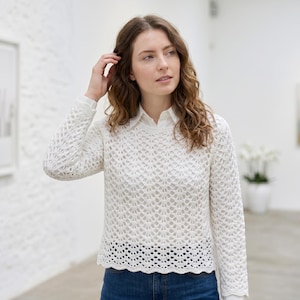 May include: A white crochet sweater with long sleeves and an openwork design. The sweater features a crew neck and a scalloped hem. A white collared shirt is worn underneath. The sweater's intricate pattern is visible.