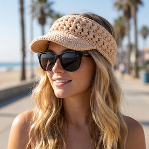 May include: A woman wearing a beige crocheted visor and black sunglasses. The visor has a textured, open weave design. The background shows a beach scene with palm trees and a sunny sky.