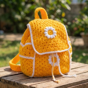 Könnte beinhalten: Ein handgefertigter gelber Häkelrucksack mit weißen Gänseblümchen-Akzenten. Der Rucksack hat einen Klappenverschluss, weiße Verzierungen und eine kleine Gänseblümchenblume auf der Vorderseite. Der Rucksack hat einen oberen Griff und verstellbare Träger. Der Rucksack steht auf einer Holzoberfläche.