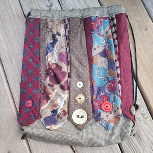 May include: A drawstring backpack constructed from vertical strips of various patterned neckties in shades of red, brown, and blue. The bag features a small black and white checkered border at the top and bottom, accented with several round buttons.