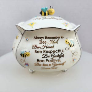 Peut inclure: Une boîte à bijoux en céramique blanche avec un couvercle orné d'une figurine d'abeille. La boîte est décorée de petites fleurs peintes, de strass bleus et d'un texte doré : Always Remember to Bee Kind, Bee Honest, Bee Respectful, Bee Grateful, Bee Positive, Bee-lieve in Yourself, I Love You!