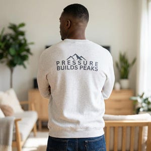 May include: A light gray sweatshirt with the text "PRESSURE BUILDS PEAKS" printed in black above a mountain graphic. The sweatshirt is a crew neck style and is being worn.