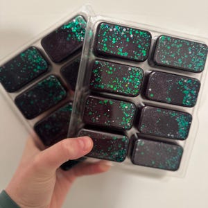 Op de afbeelding: Twee trays met rechthoekige wax melts. Elke melt is donker met groene glitter spikkels. De melts zitten in een doorzichtige plastic verpakking. De foto is genomen vanuit een iets verhoogde hoek.