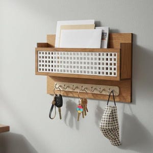 May include: Wooden wall-mounted organizer with a mail slot, key hooks, and a white grid design. The organizer holds keys, mail, and a face mask. The wood has a natural finish, and the hooks are silver.