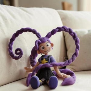 Puede incluir: Mu&ntilde;eca hecha a mano con cabello morado peinado en largas trenzas. La mu&ntilde;eca lleva una camisa morada con una estrella amarilla y una falda negra. La mu&ntilde;eca est&aacute; sentada en un sof&aacute; blanco.
