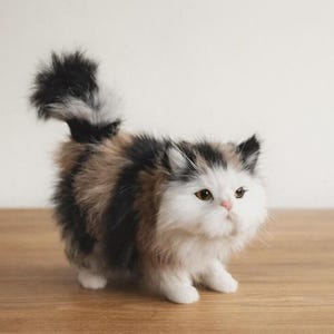 Puede incluir: Un gatito calic&oacute; de peluche, con cara blanca, ojos marrones y nariz rosa. El gatito tiene pelaje negro, marr&oacute;n y blanco y una cola esponjosa. Est&aacute; de pie sobre una superficie de madera.