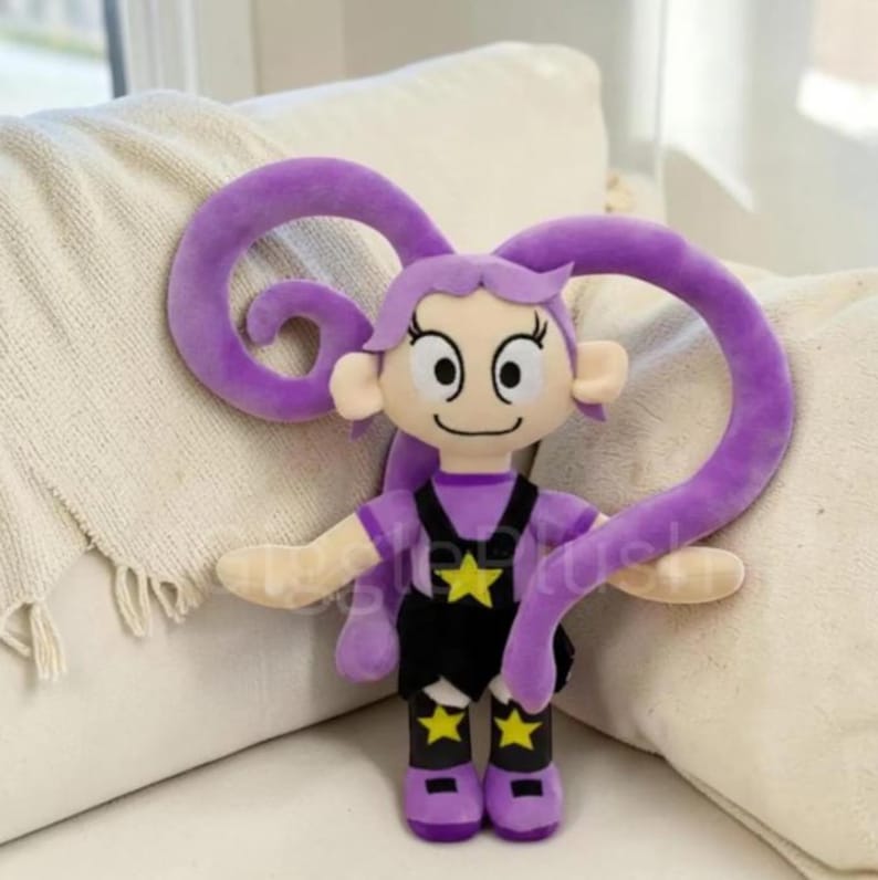 Puede incluir: Mu&ntilde;eca de peluche con dise&ntilde;o de dibujos animados. La mu&ntilde;eca tiene pelo morado, un atuendo negro con detalles de estrellas amarillas y una cara sonriente. La mu&ntilde;eca est&aacute; sobre una superficie blanca.