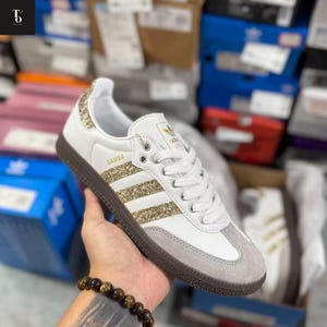 Pode incluir: Tênis Adidas Samba branco com detalhes dourados com glitter no calcanhar e nas listras. O tênis tem biqueira de camurça cinza e sola marrom. A palavra "SAMBA" está impressa na lateral.