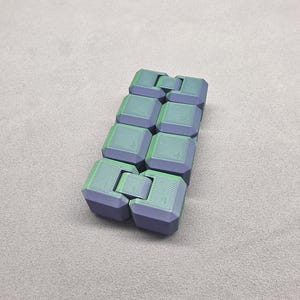 May include: A two-tone fidget toy in shades of green and purple. The toy is rectangular with eight connected blocks. Each block has a square shape with a textured surface. The toy is designed to be manipulated and played with.
