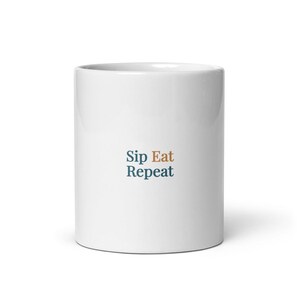 Può includere: Una tazza in ceramica bianca con il testo "Sip Eat Repeat" stampato al centro. La parola "Sip" e la parola "Repeat" sono di colore verde acqua scuro, mentre la parola "Eat" è di colore arancione tenue. La tazza ha un corpo cilindrico liscio.