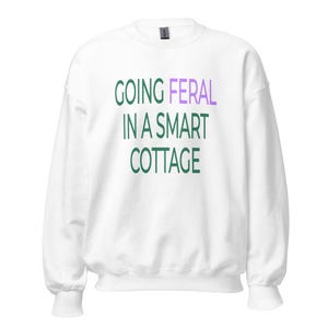 Può includere: Una felpa bianca a maniche lunghe con girocollo che presenta il testo "GOING FERAL IN A SMART COTTAGE" stampato sul davanti. La parola "FERAL" è in viola chiaro, mentre le restanti parole sono in verde scuro. Il capo ha un colletto a coste.