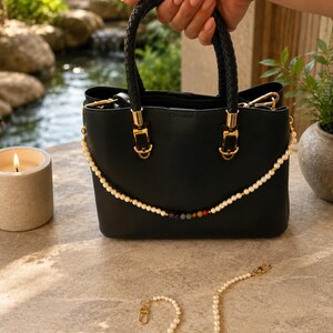 May include: Black handbag with a braided handle and gold-colored hardware. A pearl and gemstone beaded strap is attached to the bag. A separate pearl and gemstone beaded strap lies on a stone surface.