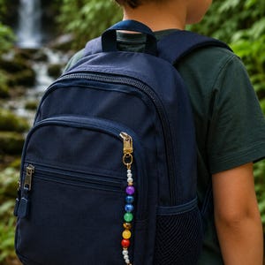 May include: Navy blue backpack with a gold zipper and a colorful beaded charm. The charm features a variety of beads in colors like purple, blue, green, yellow, and white. The backpack has a front pocket and a mesh side pocket.