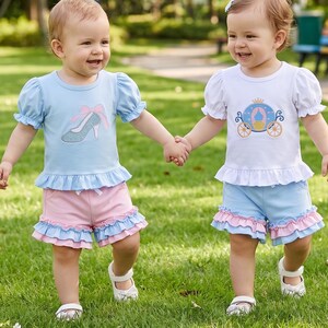 May include: Two toddlers wearing matching outfits walk on grass. One wears a light blue top with a high-heeled shoe graphic and pink shorts. The other wears a white top with a carriage graphic and blue shorts. Both outfits have ruffled hems.