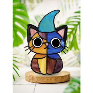 Könnte beinhalten: Eine Buntglasfigur in Form einer Katze mit blauem Spitzhut, großen runden Augen und einem mehrfarbigen Körper aus orangefarbenen, blauen, gelben und braunen Glassegmenten. Die Katze hat schwarze Drahtschnurrhaare und steht auf einem runden Holzsockel.
