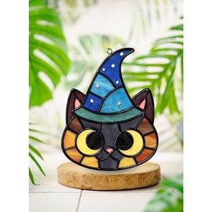 Könnte beinhalten: Ein Buntglas-Sonnenfänger in Form eines Katzenkopfes, der einen blauen Zaubererhut mit kleinen weißen Sternen trägt. Die Katze hat große gelbe Augen, eine kleine rosa Nase und ein buntes Gesicht mit Segmenten aus schwarzem, braunem und gelbem Glas.