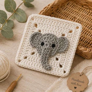 Puede incluir: Un posavasos cuadrado de ganchillo blanco roto con un aplique de elefante gris. El elefante tiene ojos negros y orejas grandes. Un ganchillo, un ovillo de lana y una etiqueta que dice "Handmade with love" también están en la imagen.