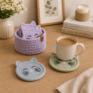May include: A set of round, crocheted cat-shaped coasters in light purple, light blue, and sage green. The coasters feature embroidered black whiskers, eyes, and a small pink nose. A matching light purple crocheted basket holds the coasters.