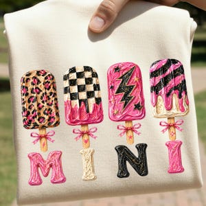 Pode incluir: Camiseta creme com quatro desenhos de picolés com vários padrões e cores, incluindo estampa de leopardo, xadrez e rosa com um raio. A palavra "MINI" está escrita abaixo dos picolés em letras decorativas.