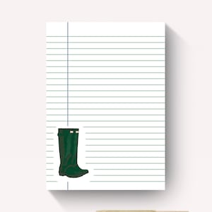 May include: A white notepad with horizontal blue lines and a vertical margin line on the left side. A small illustration of a pair of dark green rubber boots with a small white logo on the front is positioned over the lines. A brass pen is below.