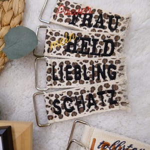 Könnte beinhalten: Nahaufnahme von mehreren Schlüsselanhängern aus Stoff mit Metallbeschlägen. Die Schlüsselanhänger haben ein Leopardenmuster und gestickte Texte in verschiedenen Farben. Die Texte enthalten Wörter wie "FRAU", "HELD", "LIEBLING" und "SCHATZ".