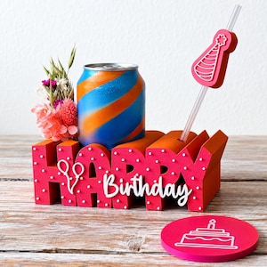 May include: A red 3D block sign spelling HAPPY with white polka dots and a white script Birthday sign. It holds a blue and orange striped beverage can, a clear straw with a pink party hat topper, and a small bouquet of dried flowers.