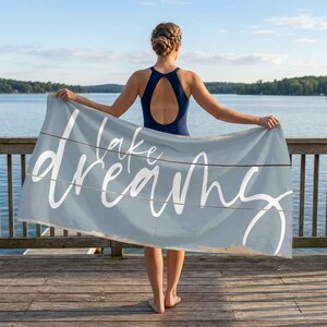 May include: A light blue beach towel with the words "Lake Dreams" in white script. The towel has a distressed, wood-plank design. The person in the image is holding the towel on a wooden dock overlooking a lake.