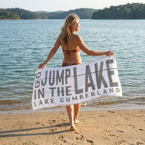 May include: A woman stands on a sandy beach holding a white towel with the text "Jump in the Lake Lake Cumberland" in gray lettering. The woman is wearing a bikini and the background shows a lake and trees.