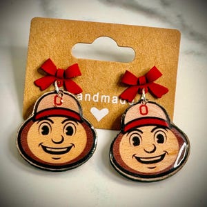 May include: A pair of dangle earrings featuring wooden charms shaped like a smiling mascot wearing a baseball cap with a red O logo. Each charm is topped with a small red bow and attached to a brown cardboard display card with the text handmade.
