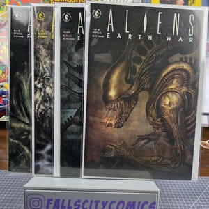May include: A collection of four Aliens: Earth War comic books. The first comic features a detailed illustration of an alien creature. The comic books are displayed on a gray stand with the text "FALLSCITYCOMICS".