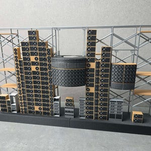 May include: A miniature model of a server farm featuring two tall towers of stacked black computer fans and server components, a large cylindrical unit, and a smaller rectangular unit, all supported by a silver metal scaffolding frame on a black base.