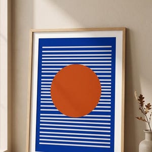 Puede incluir: Una lámina de arte mural rectangular enmarcada que presenta un círculo naranja sólido centrado sobre una serie de líneas blancas horizontales sobre un fondo azul intenso. La obra está en un marco de madera clara con borde blanco.