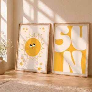 Puede incluir: Dos láminas verticales enmarcadas. La lámina de la izquierda muestra un sol amarillo con cara y pequeñas estrellas sobre un fondo crema. La lámina de la derecha muestra la palabra SUN en letras blancas grandes y gruesas sobre fondo amarillo.