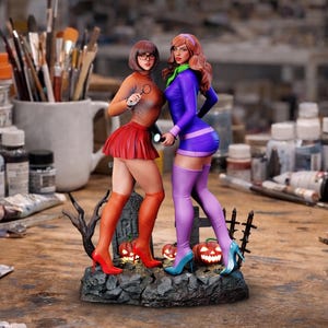 Peut inclure: Figurine d'Halloween représentant deux personnages féminins costumés. L'une porte un haut orange, une jupe rouge et des lunettes, tenant une loupe. L'autre porte une robe et des bottes violettes. Elles se tiennent sur une base avec des citrouilles et une pierre tombale.
