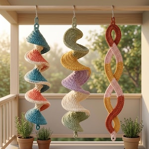 Könnte beinhalten: Drei hängende Häkelornamente, die an einer Holzstange befestigt sind. Das linke Ornament ist eine Spirale in Blaugrün und Koralle. Das mittlere Ornament ist eine Spirale in Salbeigrün, Gelb und Lavendel. Das rechte Ornament ist eine geflochtene Schlaufe in Rost, Rosa und Gelb.
